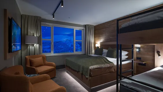 A cozy hotel room features a large bed and bunk beds, wooden walls, and orange armchairs. A window reveals a snowy mountain landscape under a blue sky. A television is mounted on the wall.