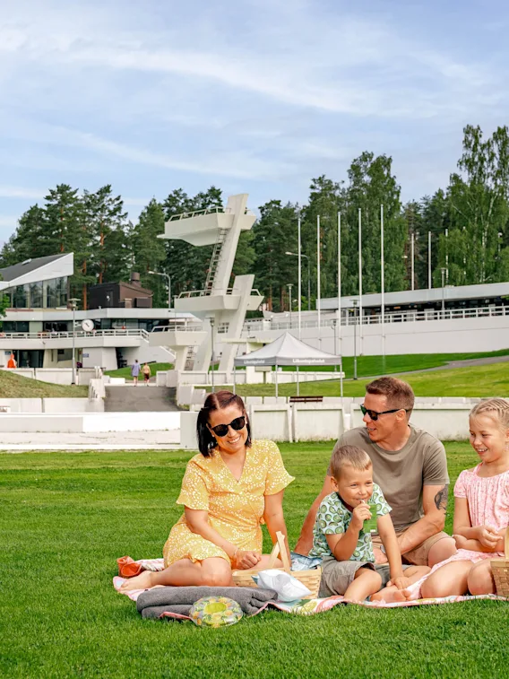 Perhe istuu piknikhuovalla, syö ja keskustelee. Heitä ympäröi laaja vihreä nurmikko, jossa on puita ja moderneja rakennuksia taustalla.