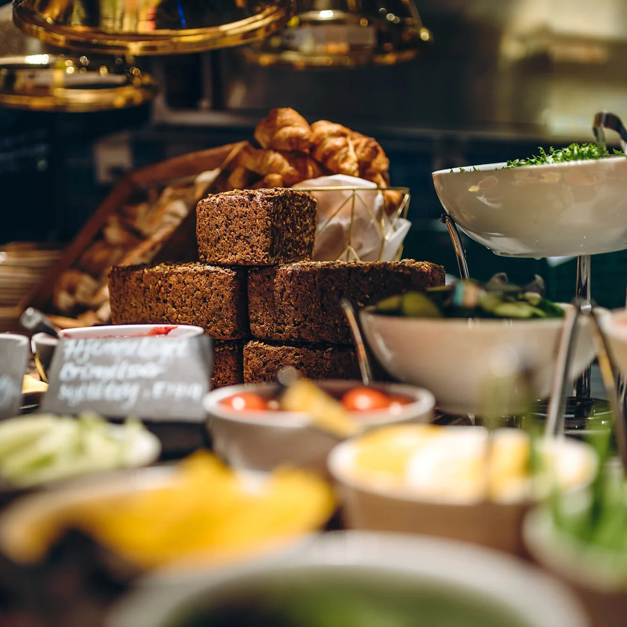Bread slices stacked on a buffet table surrounded by dishes of salad and toppings. Labels read “Hjemmelavet Mini-Knoldsellerisalat Etnis” and “Hjemmelavet Grønkålssalat.”