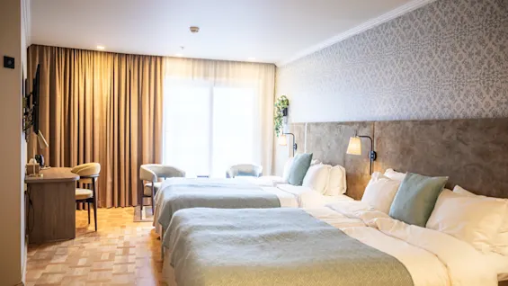 Två prydligt bäddade dubbelsängar med ljus sängkläder och blå kuddar i ett mysigt hotellrum med mönstrad tapet, ett litet skrivbord och ett stort fönster med fördragna gardiner.