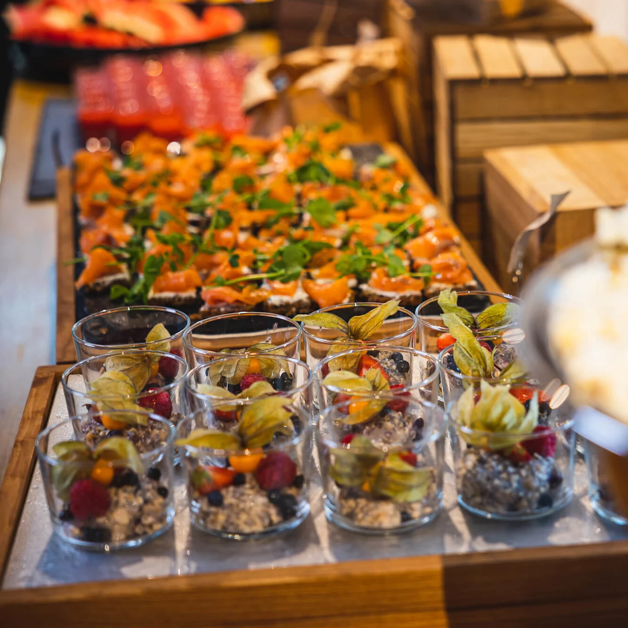 Glass fylt med bær og granola står på et trebord ved siden av appetittvekker toppet med grønnsaker og trekasser i bakgrunnen.