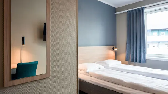 A bed with white sheets and pillows stands in a minimalistic room with wooden floors and a window with blue curtains; a mirror and lamp are visible on the left wall.