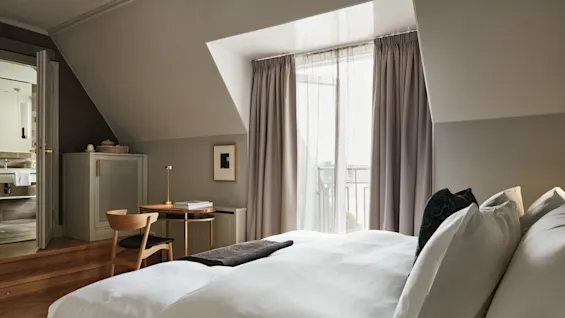 Seng med hvitt sengetøy ligger i et moderne soverom, ved siden av et lite skrivebord i tre med en stol. Gardiner dekker et stort vindu med en balkong, ved siden av et bad.