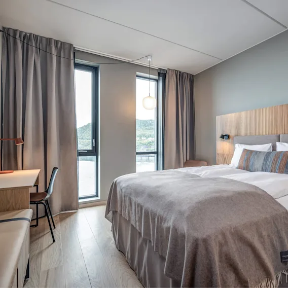 En pent oppredd seng med grått pledd og puter står mot en trevegg. Den står i et moderne rom med skrivebord, stol og stort vindu.