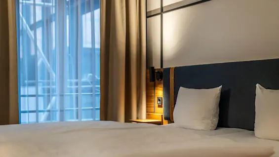  A close-up of a neatly made double bed with white bedding. The headboard is upholstered in blue fabric with wooden panel details. Behind the bed is a bedside table with a reading lamp, and large windows with beige curtains let in soft light.