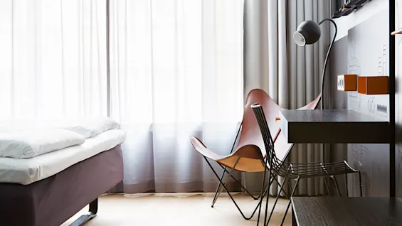 Seng med hvitt sengetøy står ved siden av en moderne stol og skrivebord med lampe i et rom med gjennomsiktige gardiner.
