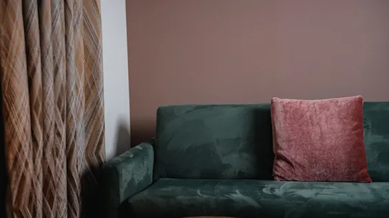 En komfortabel grønn fløyelssofa med en rosa pute, ved siden av et mønstret gardin, som skaper en koselig atmosfære.