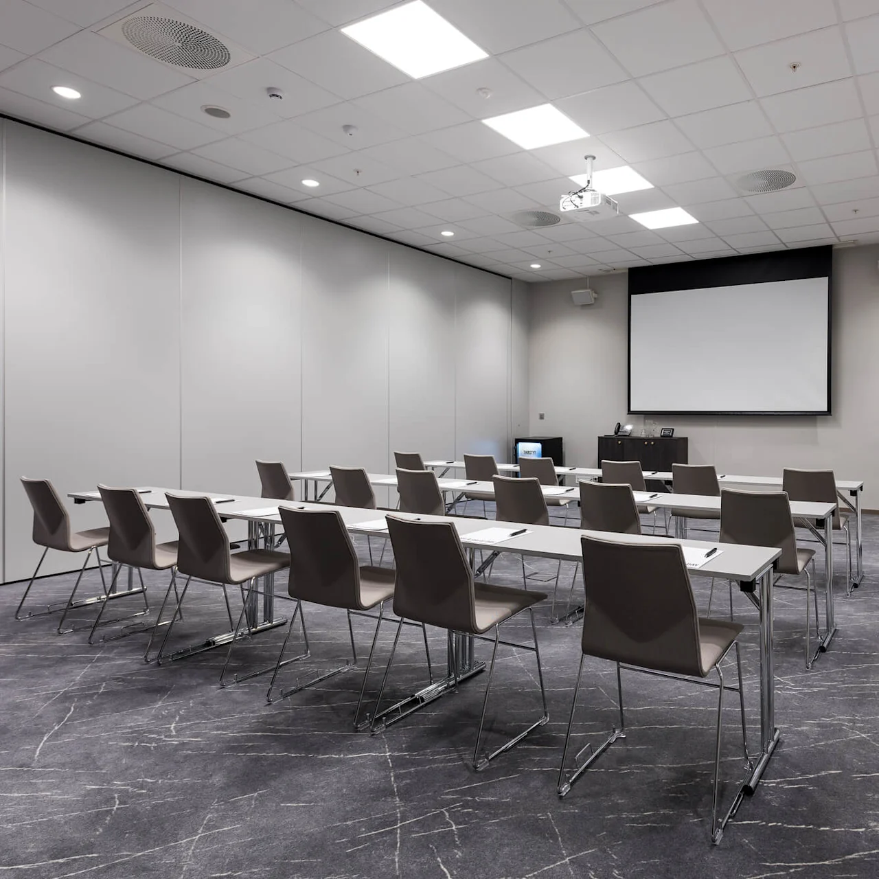 Rader av tomme stoler og bord vender mot en tom projektorskjerm, plassert i et moderne konferanserom med grått teppe og en whiteboard på høyre vegg.