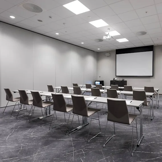 Rader av tomme stoler og bord vender mot en tom projektorskjerm, plassert i et moderne konferanserom med grått teppe og en whiteboard på høyre vegg.