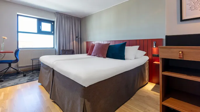 En pent oppredd seng dominerer et moderne hotellrom med enkel innredning, kunst på veggen og dagslys som strømmer inn gjennom et vindu.