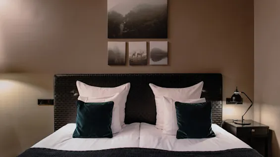 Et koselig hotellrom med en pent oppredd seng, mørkegrønne puter og naturkunst på veggen, som skaper en fredelig atmosfære.