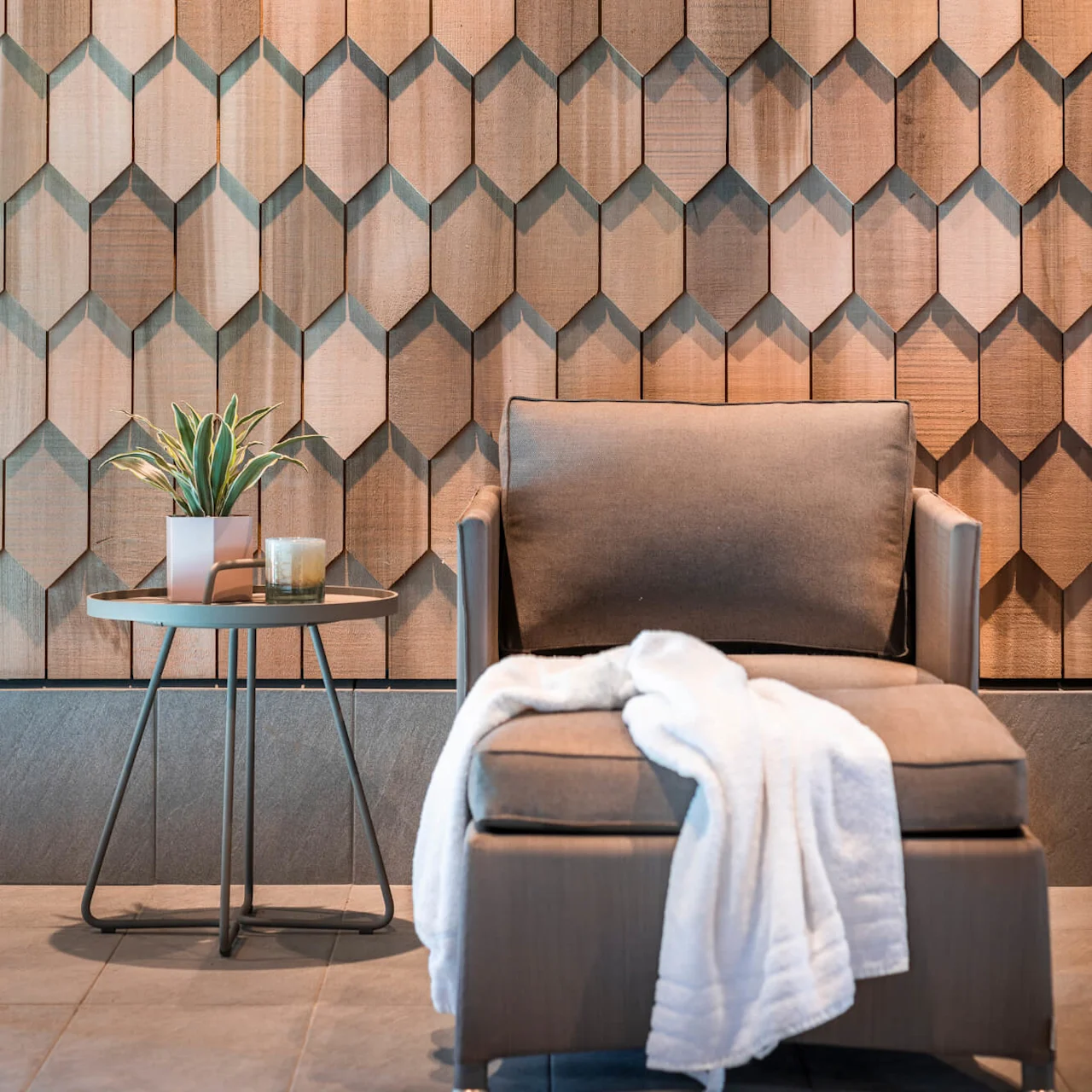 A brown armchair with a white blanket rests against a textured wood-paneled wall, next to a small round table holding a plant and candle on a tiled floor.