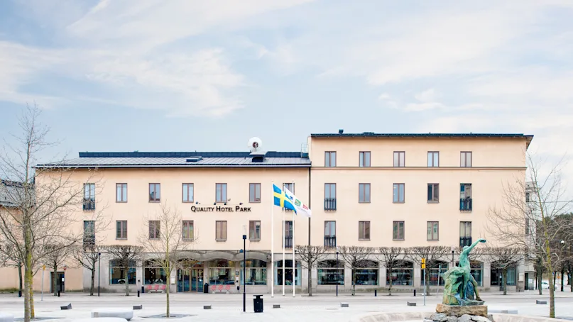 A tan-colored building labeled "Quality Hotel Park" stands behind a paved square. A statue on a stone platform and two flagpoles with Swedish flags are in front, among leafless trees.
