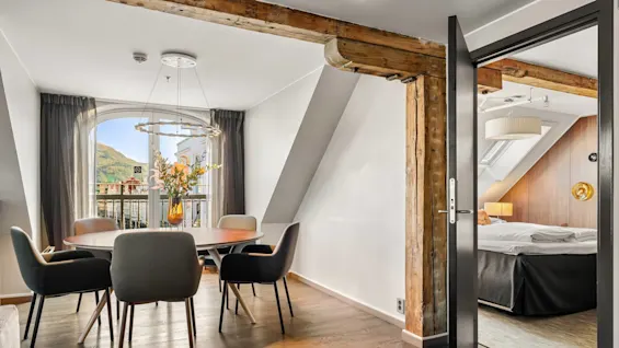 Et rundt spisebord med fire stole står ved et stort vindue med bjergudsigt, under en moderne pendellampe. Synlige træbjælker adskiller spiseområdet fra et soveværelse.