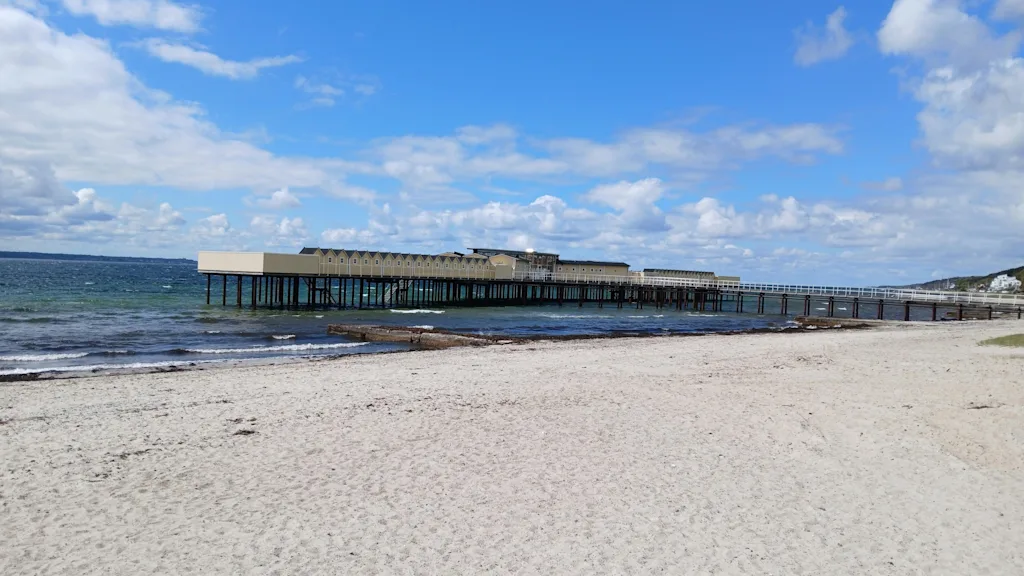 En lang brygge med bygninger strekker seg ut i havet fra en sandstrand under en blå himmel med hvite skyer.