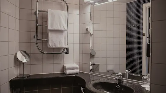 Siisti ja moderni kylpyhuone Home Hotel Uppsalassa, jossa on tumma pesuallas, suuri peili ja raikkaita valkoisia pyyhkeitä lämmitetyllä telineellä.