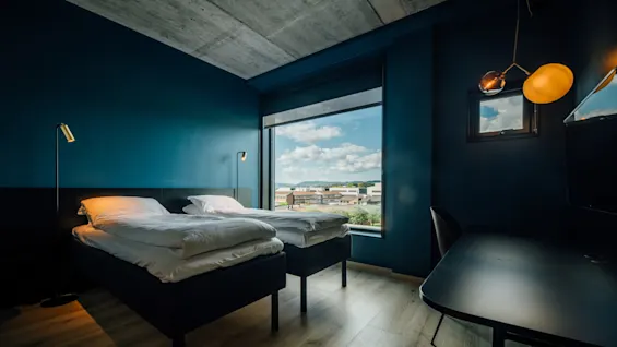 Et moderne hotelværelse med to sirligt redte senge og stilige lamper, med store vinduer der viser et bylandskab under en blå himmel.