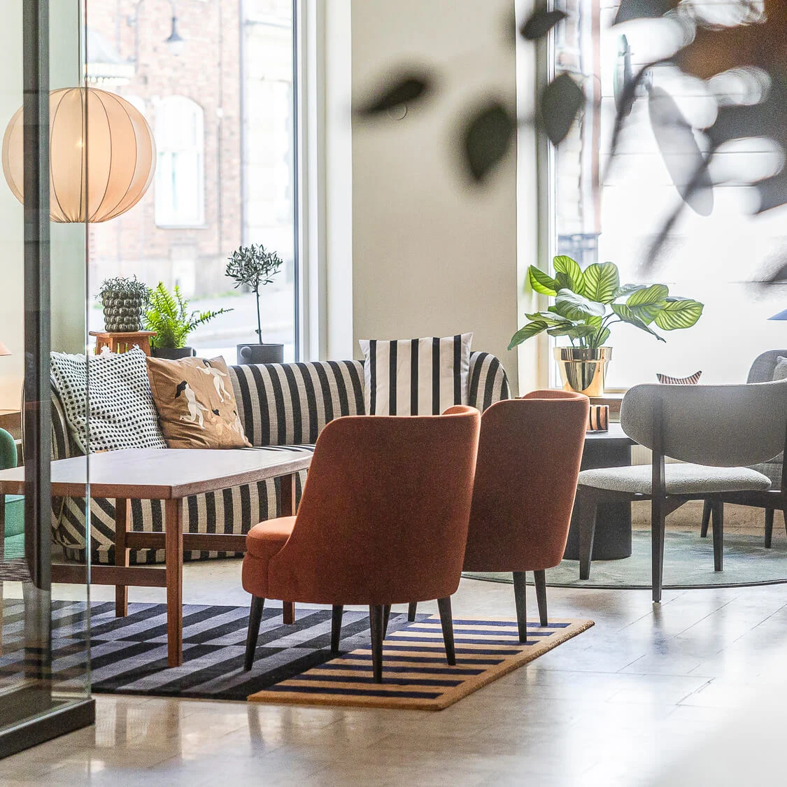 En modern lounge med stilfulla stolar och soffor, växter och en mönstrad matta, sedd genom en glasvägg.