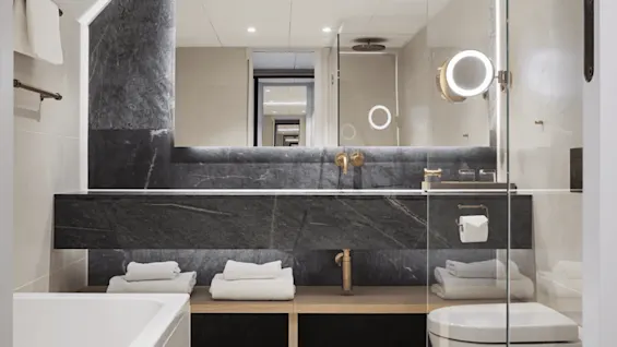 A modern bathroom features a large mirror, illuminated by spotlights, reflecting a continuous hallway. Towels are neatly placed under a dark stone countertop beside a bathtub.