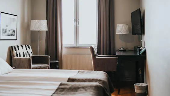 Et hyggeligt soveværelse med redt seng, to lænestole, lamper og et tv, belyst af naturligt lys fra et vindue med gardiner.