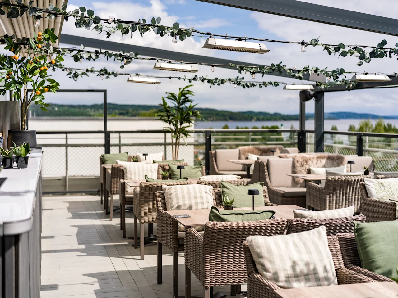 Wickerstolar med kuddar står på en takterrass, under en pergola dekorerad med lampor och grönska. Bakgrunden visar en naturskön utsikt över avlägsna träd och vatten.