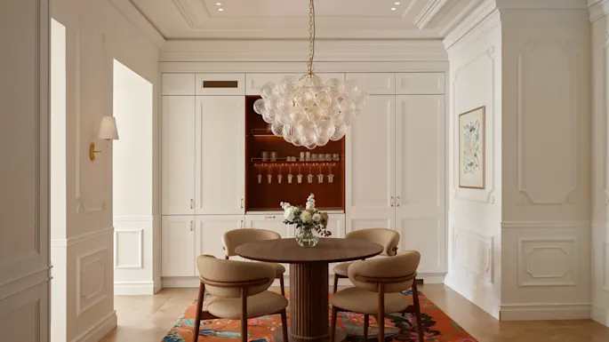 Pyöreä puupöytä tuoleineen seisoo lasisen kattokruunun alla tyylikkäässä ruokailuhuoneessa, jossa on valkoiset paneeliseinät, sisäänrakennetut kaapit, kukkataidetta ja värikäs kuviomatot.