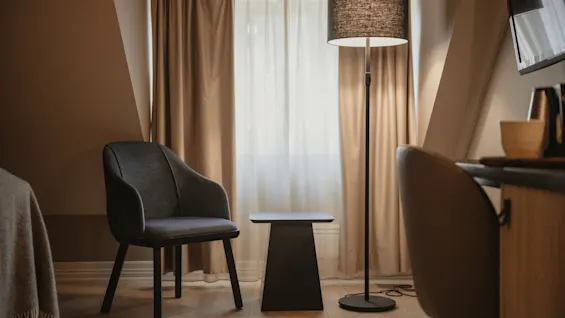 Et hyggeligt hotelværelse med en komfortabel stol, et lille bord og en gulvlampe ved et vindue med gardiner, der skaber en varm atmosfære.
