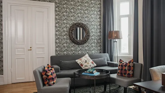 En grå sofa og stole med farverige puder står omkring et sort sofabord i en stue med blomstrende tapet og et rundt spejl. Store vinduer giver naturligt lys.