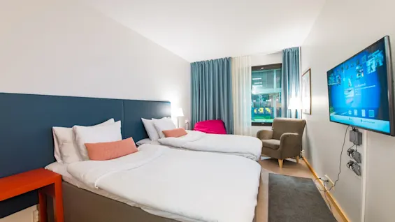 A hotel room with two neatly made beds, featuring white linens and orange cushions. A television is mounted on the wall, opposite an armchair near a window; a small red table stands beside the beds.