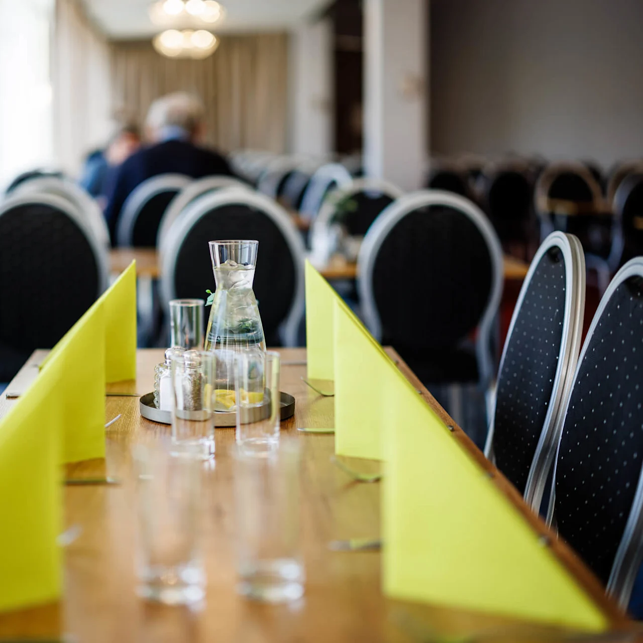 Ett långt matbord med gröna vikta servetter och en glaskaraff med vatten och citronskivor. Tomma stolar omger bordet i en väl upplyst restaurangmiljö.