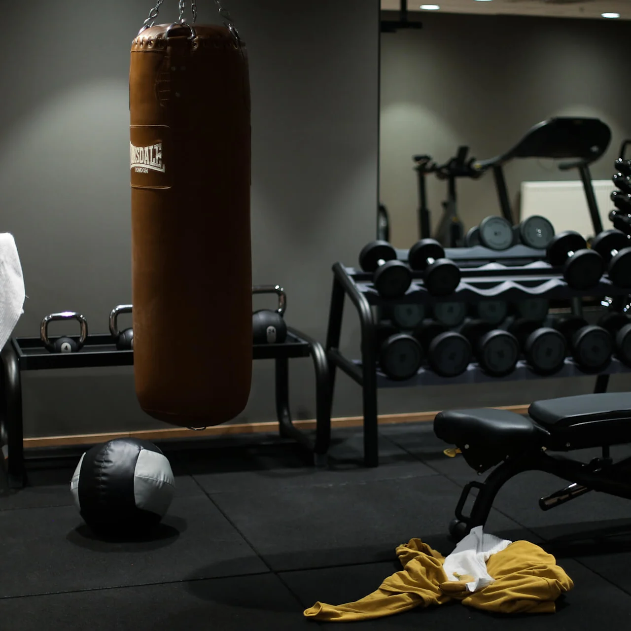 Punching bag hangs in gym with weights and bench nearby; towel and yellow jacket on floor. Text on bag: "Lonsdale London". Mirrors, dumbbells, and exercise machines fill the space.