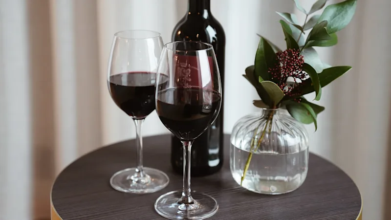 To glas rødvin og en flaske på et bord, med en vase der indeholder grønt og bær.