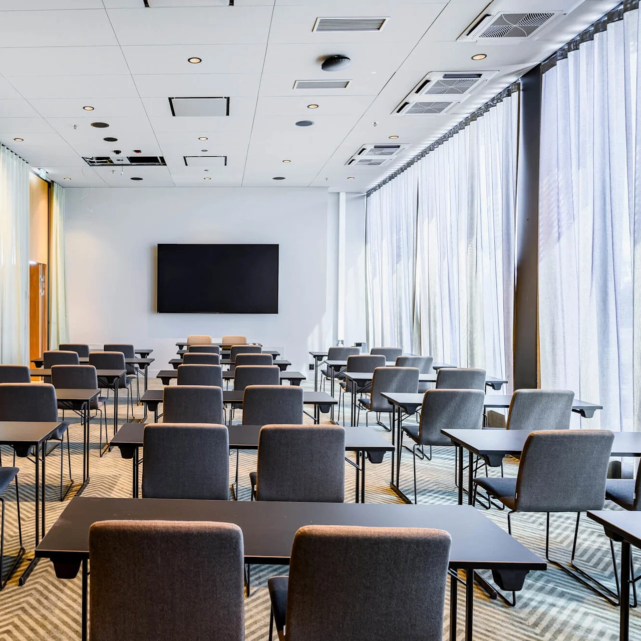 Rows of gray chairs and rectangular tables face a large blank screen in a bright conference room. Soft lighting streams through sheer curtains along tall windows.