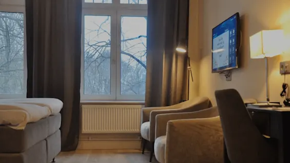 Hotellihuone, jossa sänky, tuolit, pöytä ja televisio, suuren ikkunan kehystämänä paljaita puita.