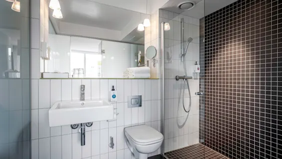 En vit handfat är monterat på en kaklad vägg, bredvid en toalett och en glasomsluten dusch. Handdukar staplade på en hylla ovanför, upplysta av taklampor.