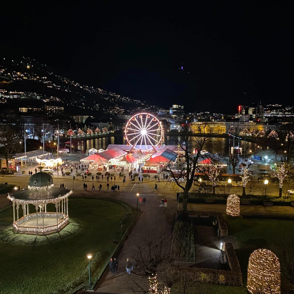 A festive night scene with a brightly lit Ferris wheel, a gazebo, and a bustling market area with many people.