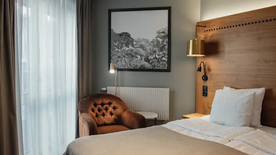 Et moderne hotelværelse med en komfortabel seng, en plysj lænestol og et stort vindue med gardiner, der skaber en afslappende atmosfære.