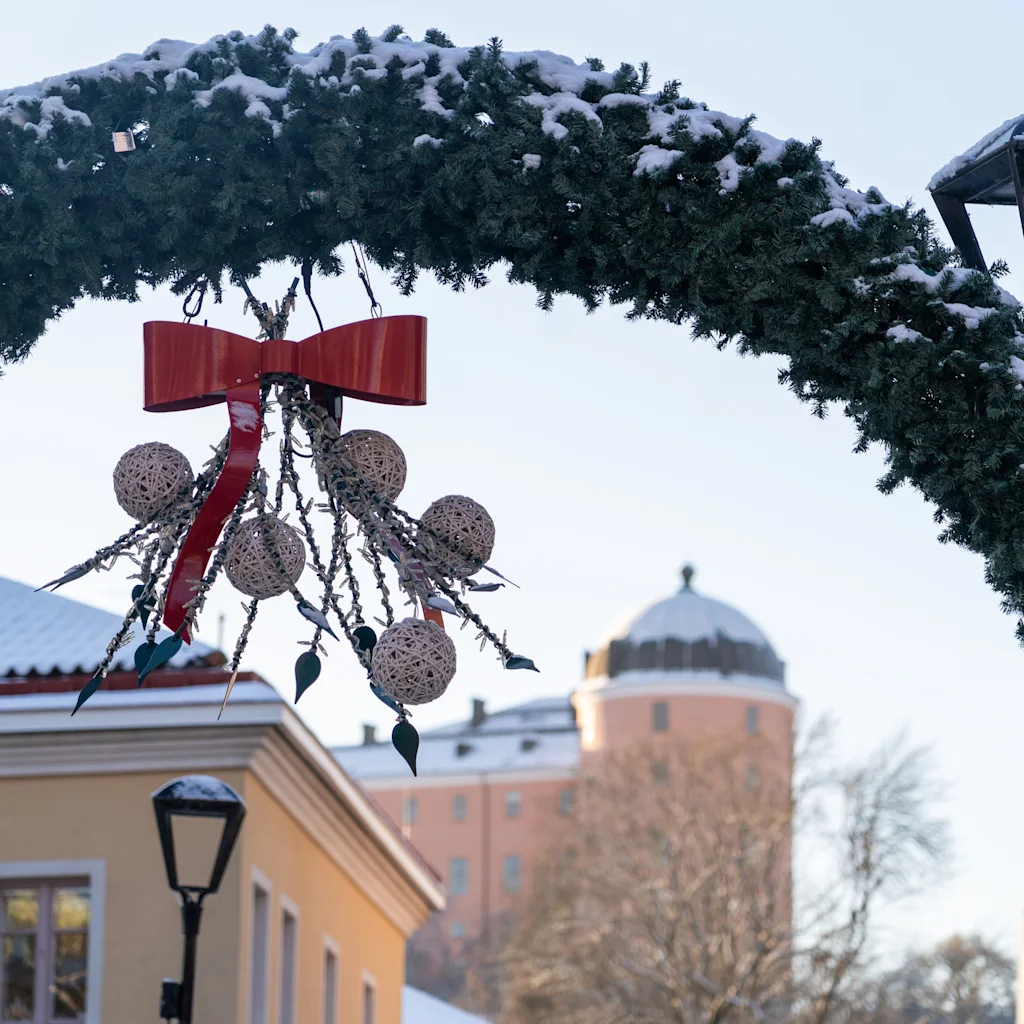 A snow-covered evergreen arch with a red bow and ornaments decorates a winter street scene, with buildings and a sign reading KYRKAN.