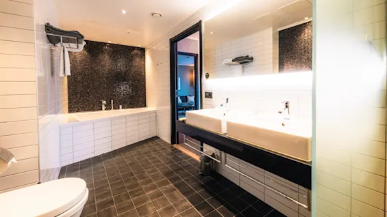 Badekar er stille på badeværelset, omgivet af hvide fliser. Dobbelt vaske vender mod et stort spejl med pænt arrangerede håndklæder. En dør åbner ud til et tilstødende rum. Moderne, elegant atmosfære.
