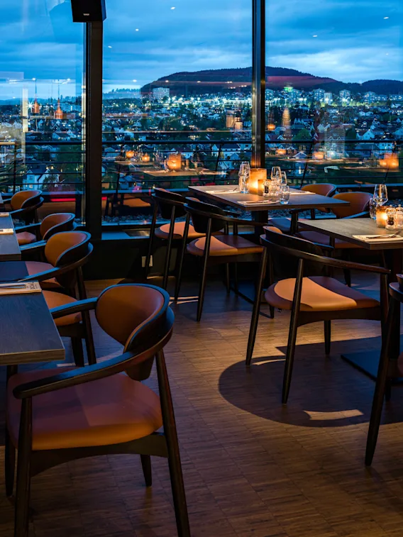 Mysig restaurang med dukade bord och levende ljus framför stora fönster med utsikt över staden i skymningen.