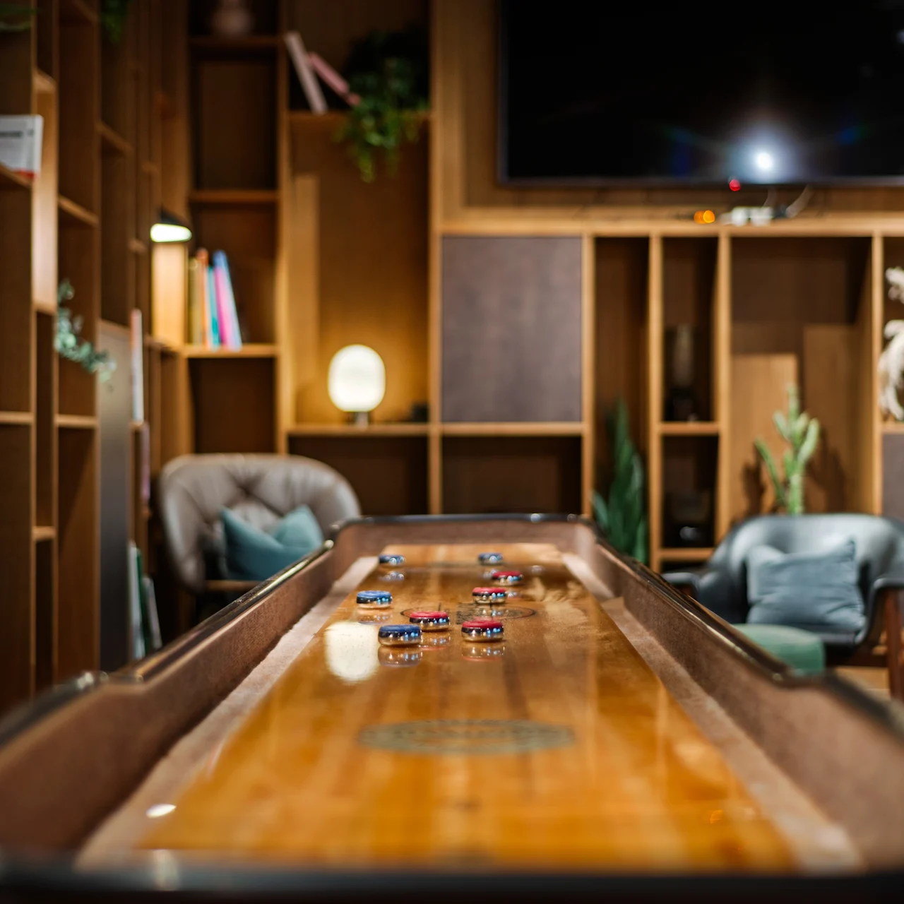 Et shuffleboardbord i en hyggelig lounge med skiver i spil, omgivet af træhylder, planter, lænestole og dæmpet belysning, der skaber en afslappet atmosfære.