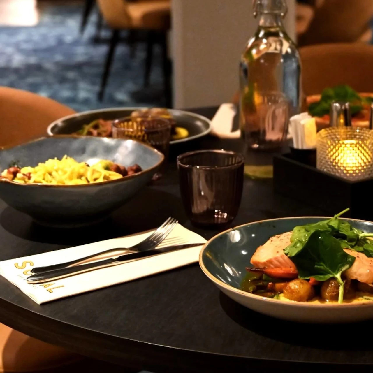 En matsalsbord med tallrikar med lax, grönsaker och pasta, upplyst av ett litet ljus. En flaska, tomma glas och bestick på en servett med texten "SUAL" kompletterar bilden.