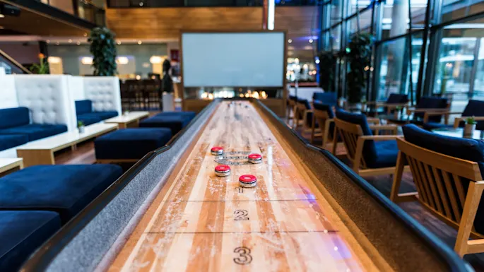 A shuffleboard table with red pucks is positioned in a modern lounge, surrounded by blue cushioned seating, tables, and large windows.  