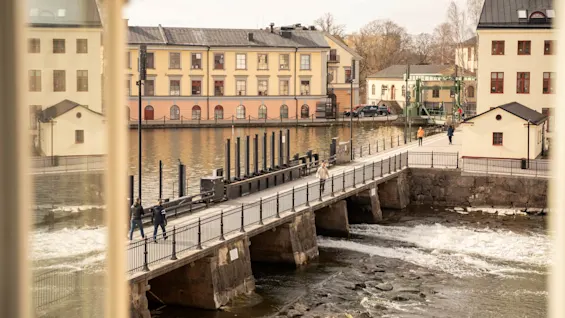 En bro sträcker sig över en flödande flod, med människor som går över. Omkring finns historiska byggnader med rödprydda fönster. Scenen ramas in av två oskarpa fönsterkanter i förgrunden.