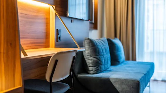 A modern hotel room featuring a wooden desk illuminated by a built-in LED light and a simple chair. Next to it is a comfortable sofa with blue cushions, and large windows with light curtains are visible in the background.