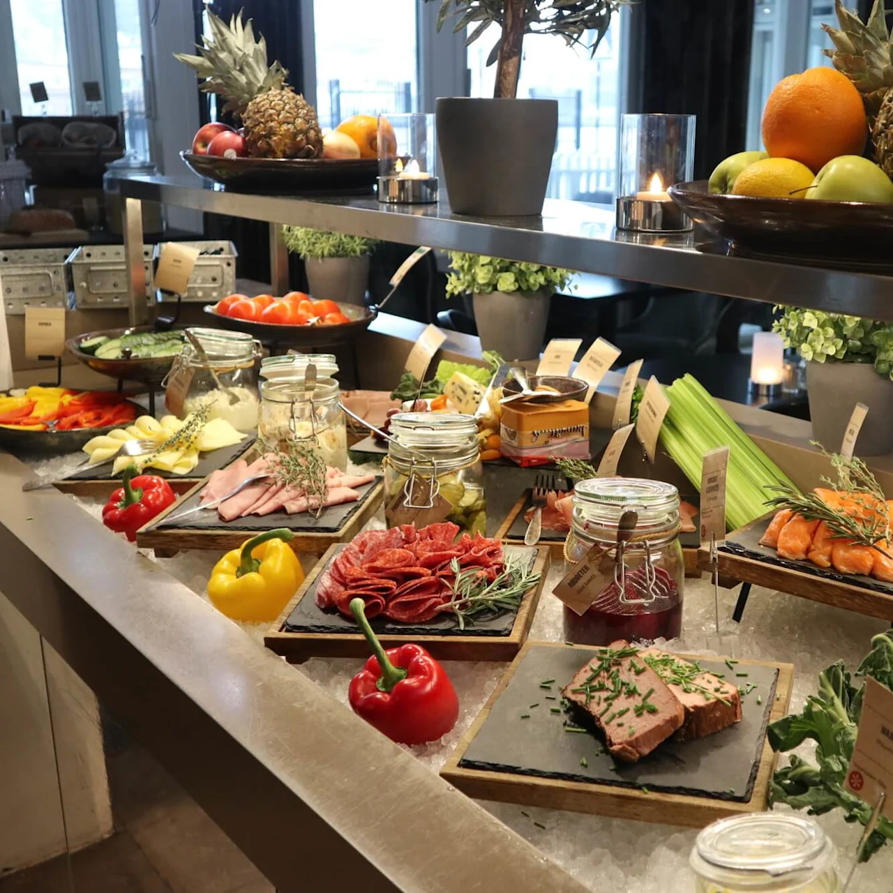 A variety of foods are displayed on a buffet with labels, including fruits, cheeses, meats, and condiments, in a well-lit room.