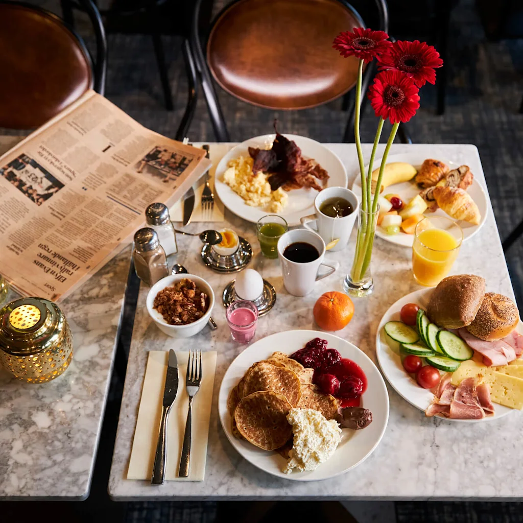 Breakfast table set with pancakes, eggs, bacon, fruits, cheese, ham, croissants, coffee, and juice. Red flowers in a vase and an opened newspaper add to the dining atmosphere.
