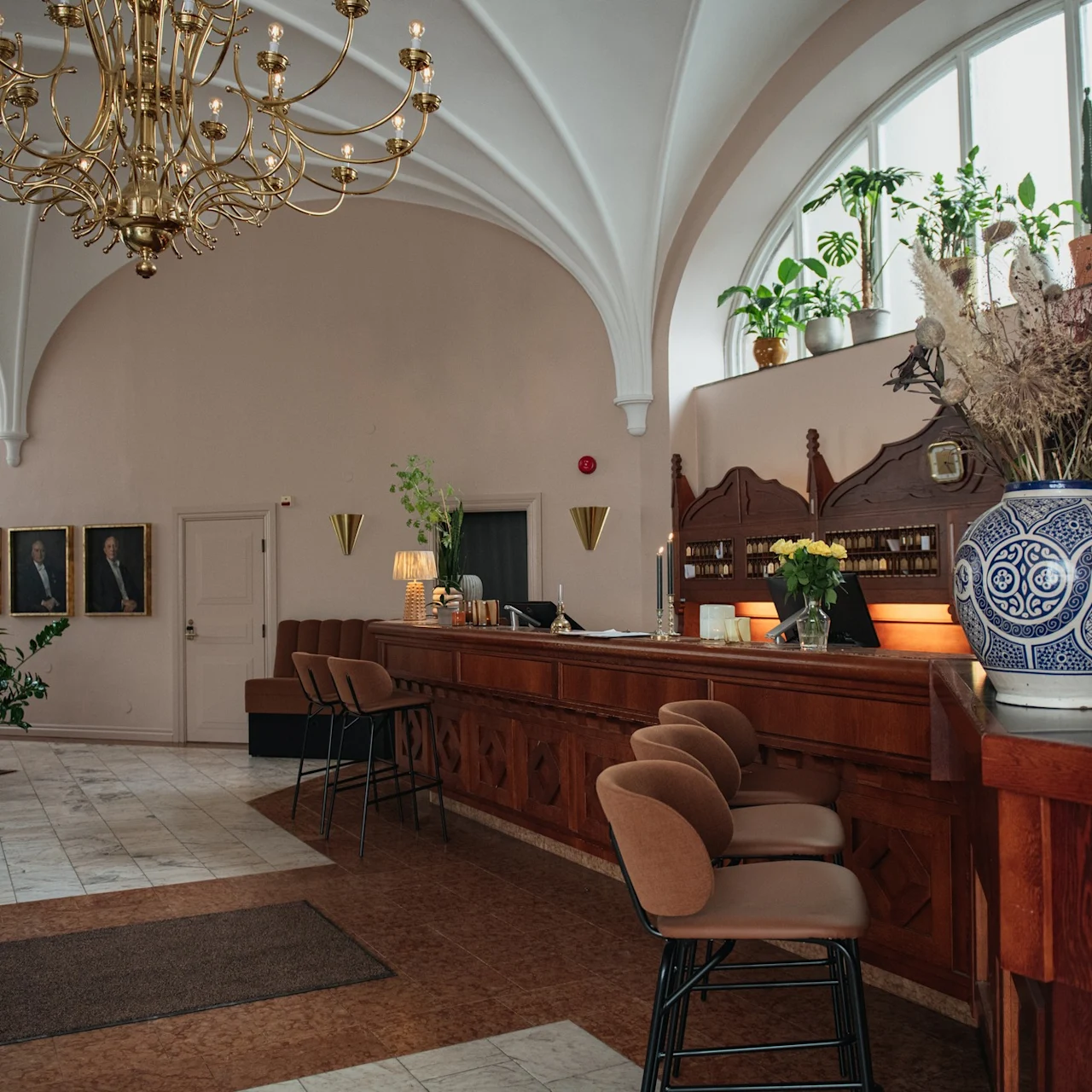 A chandelier hangs above a hotel lobby with a large wooden bar. Chairs line the bar, and plants sit on top of shelves, adding greenery to the elegant room.