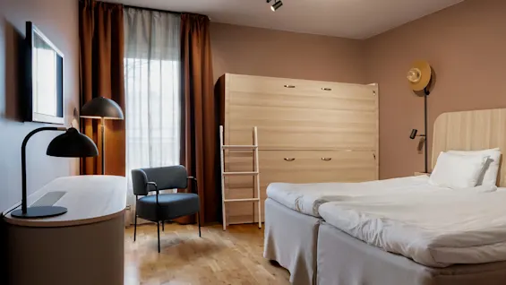 Ett mysigt hotellrum med två enkelsängar, ett skrivbord, en fåtölj och en smart väggenhet med stege.