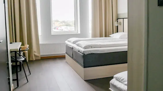 En seng er pent oppredd i et lite, lyst rom med et stort vindu. Beige gardiner og et skrivebord med stoler er i nærheten.